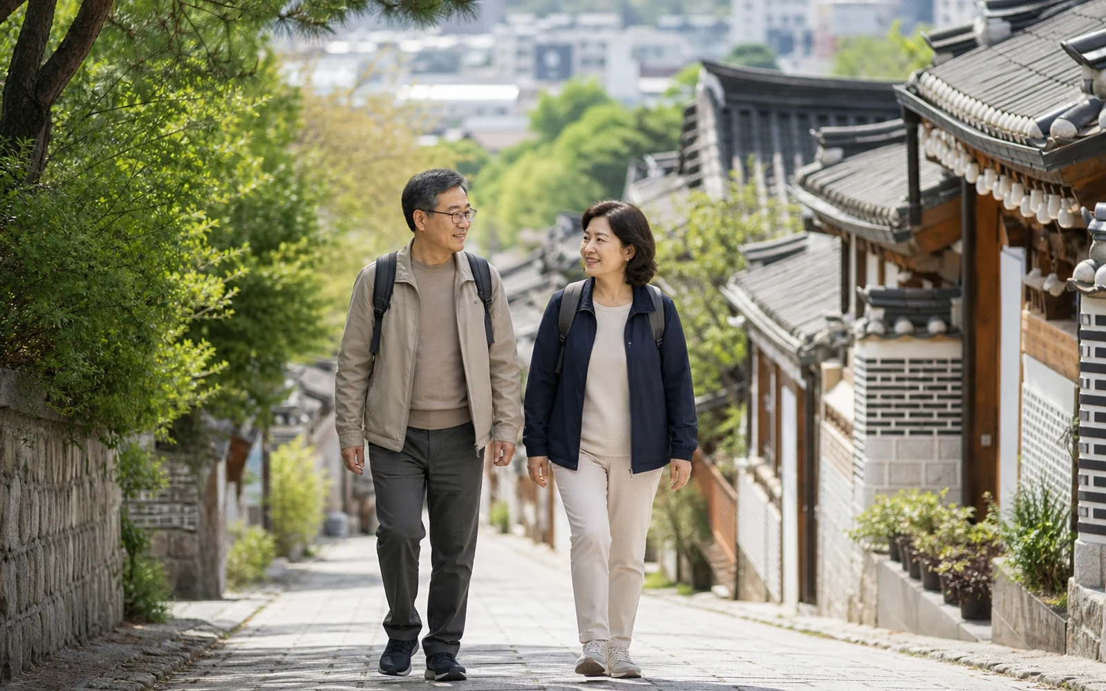작은 배낭을 메고 전통 지붕이 보이는 조용한 마을길을 걷는 50대 부부